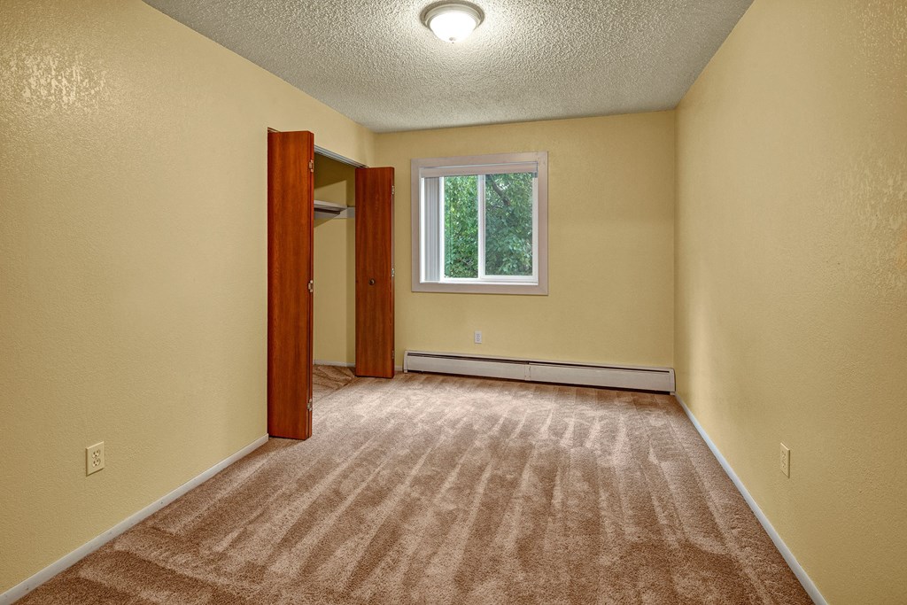 Empty bedroom with yellow walls, beige carpet, and a single window showing greenery. There’s an open closet with wooden doors on the left. Cozy and simple. Arctic Sun Apartments.