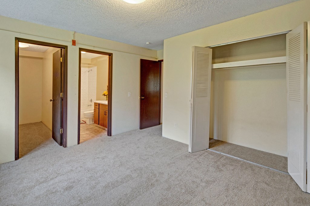 Empty bedroom with beige carpet, open closet with white doors on the right, and two wooden doors on the left leading to a bathroom and another room. Arctic Sun Apartments.