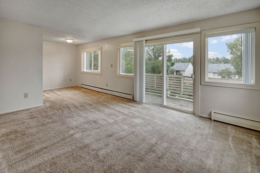 Spacious carpeted living room with large windows and sliding glass door leading to a balcony. Light gray walls and ceiling create a bright, open feel. Arctic Sun Apartments.