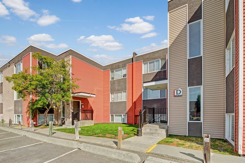 Arctic Sun Apartments building with red and beige siding, two stories, and a "D" sign for units 288-350. Trees and parking spaces in the foreground. Sky is clear with scattered clouds.