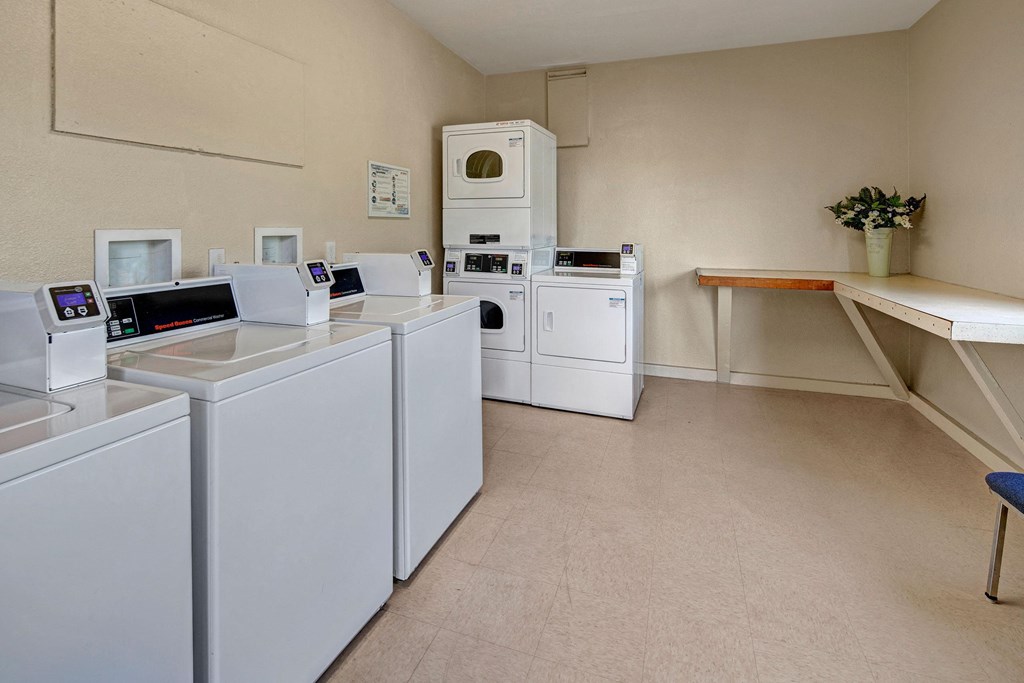A clean laundry room here at Bay Arms Apartments with three white washing machines and two stacked dryers. A folding table with a vase of flowers adds a welcoming touch.
