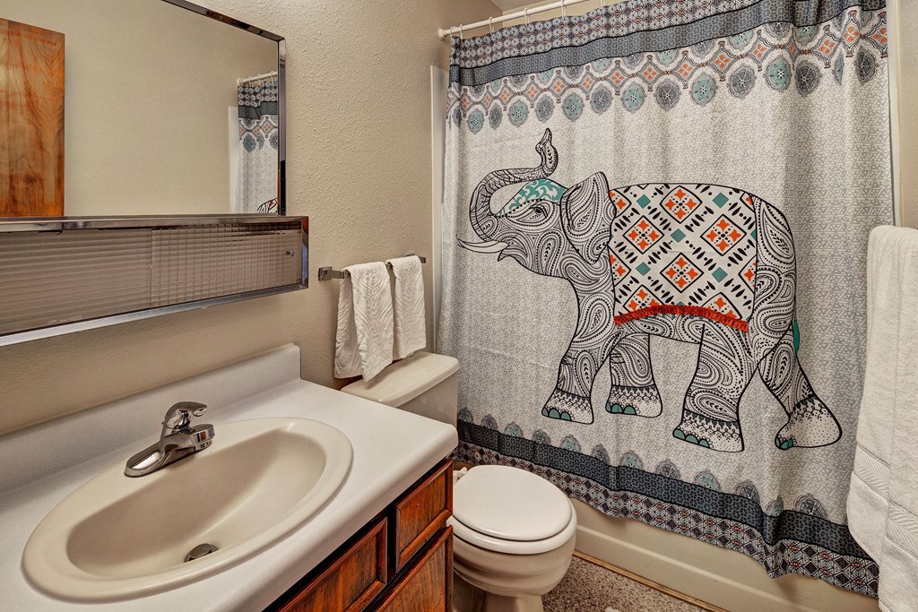 A bathroom here at Bay Arms Apartments with a patterned elephant shower curtain. Features a white sink, wooden cabinets, a mirror, and neatly hung white towels, conveying a cozy atmosphere.