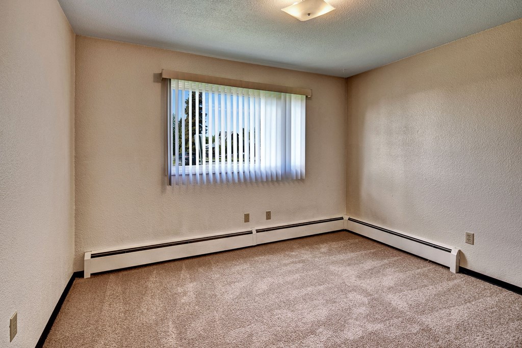 Empty beige bedroom here at Bay Arms Apartments with carpeted floor, a bright window with vertical blinds, and baseboard heating. Soft overhead light creates a calm atmosphere.