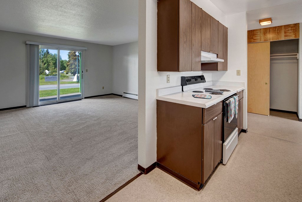 Small kitchen here at Bay Arms Apartments and empty living room with beige carpet. Kitchen features dark wood cabinets and an electric stove. Large sliding glass doors lead outside, creating a bright, open atmosphere.