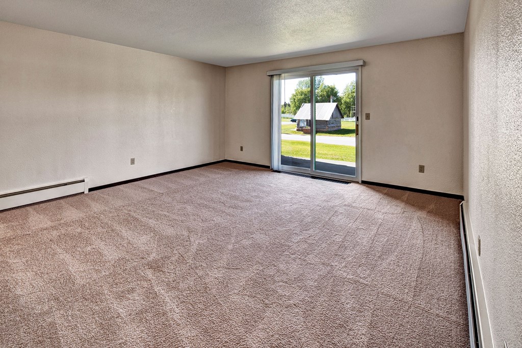 Empty living room here at Bay Arms Apartments with light brown carpet and beige walls, featuring a large sliding glass door opening to a grassy yard. Sunlight illuminates the space.