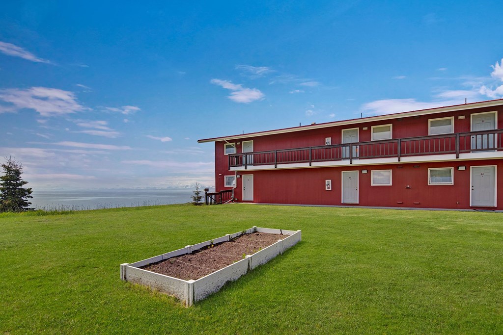 Our red barn-style building here at Bay Arms Apartments with multiple doors and windows sits on a lush green lawn. A white raised garden bed lies in front. The sky is clear and blue.