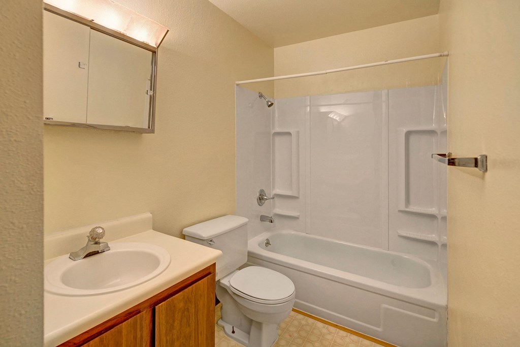 Brighton Woods Apartments - Bathroom Compact beige bathroom here at Brighton Woods Apartments with a white bathtub, showerhead, toilet, and sink with a mirror above. Simple, clean, and functional design.