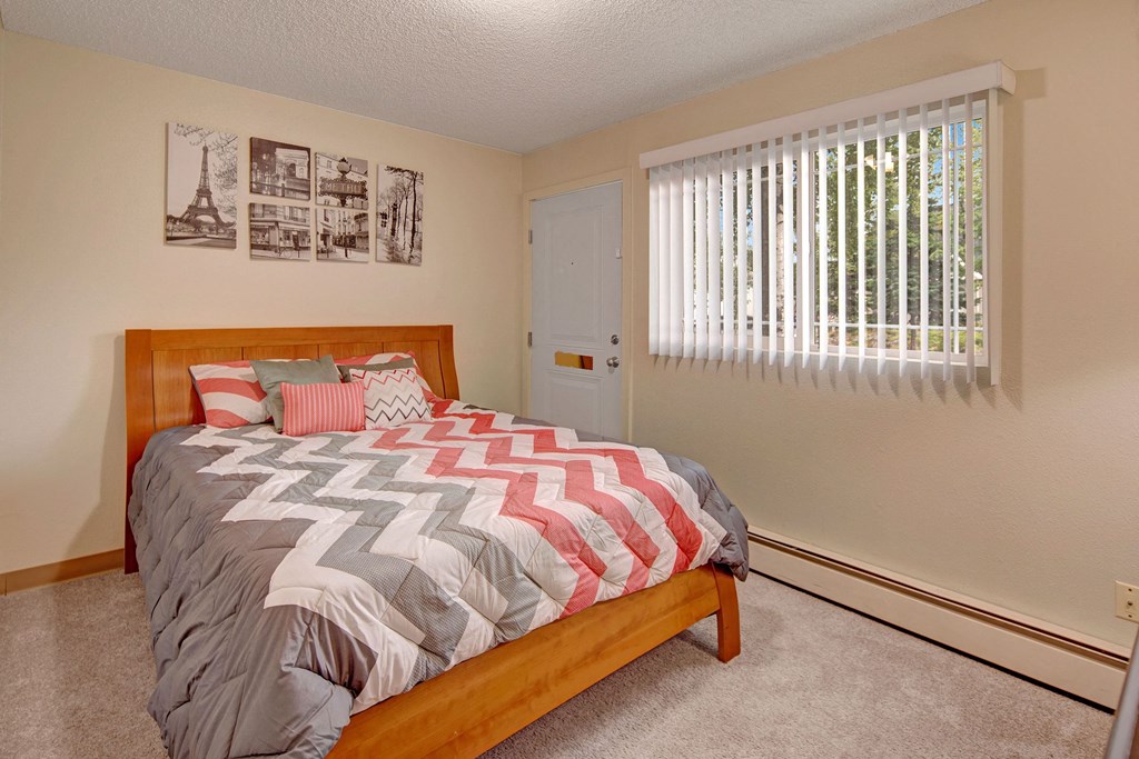 Brighton Woods Apartments - Bedroom Cozy bedroom here at Brighton Woods Apartments with a wooden bed, chevron-patterned bedding in gray, white, and pink. Sunlight filters through vertical blinds on a window, brightening the space. Black and white art above the bed adds a modern touch.