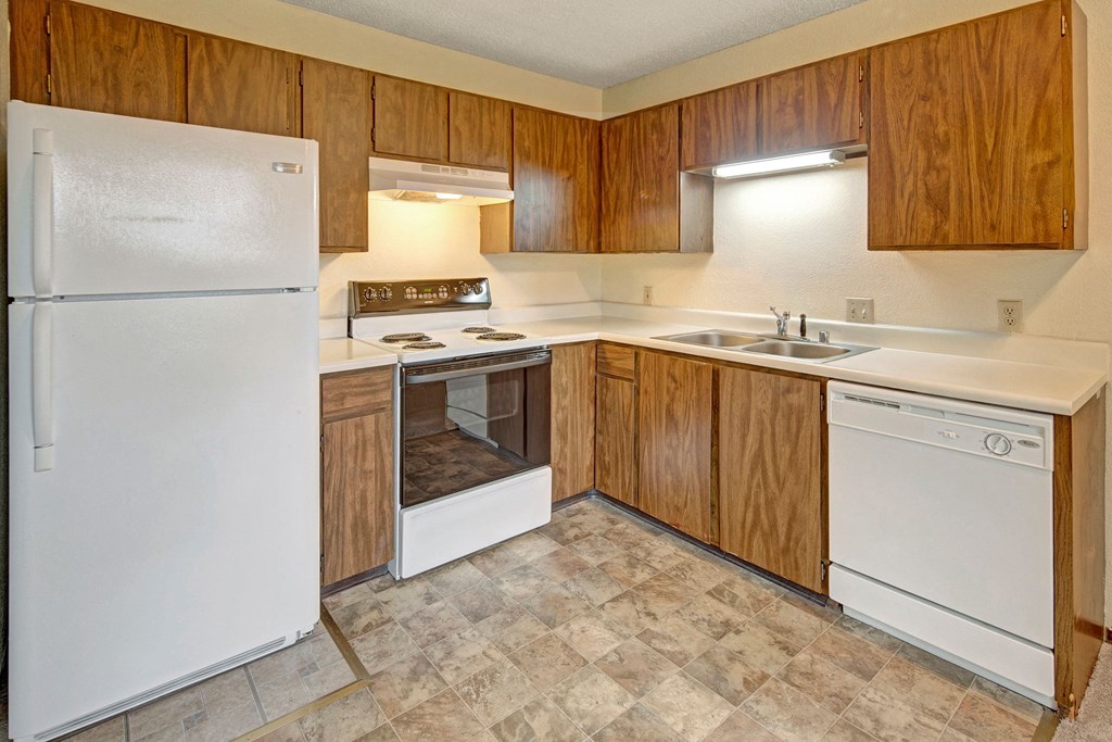 Simple kitchen here at Brighton Woods Apartments with wooden cabinets, white appliances including a fridge, stove, and dishwasher. Neutral tones create a clean, functional space.