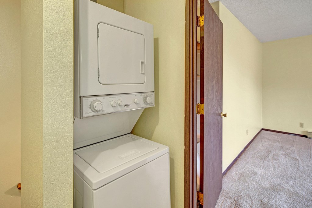 Stacked washer and dryer here at Brighton Woods Apartments in a small laundry area with beige walls. An open wooden door leads to a carpeted room, creating a cozy and tidy atmosphere.