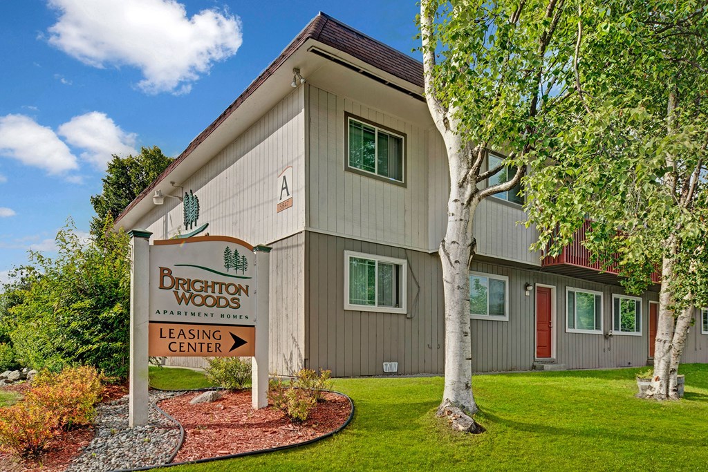Brighton Woods Apartments - Exterior Apartment complex with a beige facade and red door, surrounded by lush greenery and birch trees. A sign reads "Brighton Woods Leasing Center."
