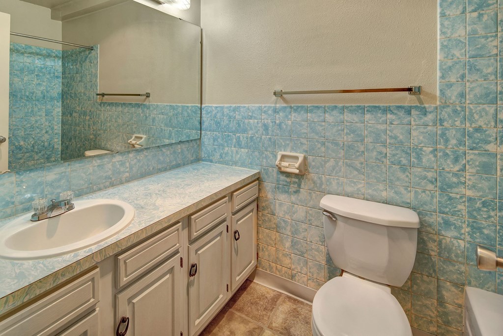Bathroom here at Castle Apartments with light blue, textured tile walls, a large mirror, and white fixtures. Features a white sink and toilet against a beige floor.