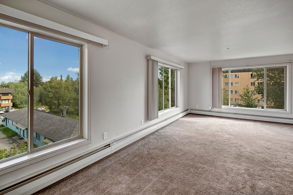 Living room here at Castle Apartments with large windows on two walls, beige carpet, and white walls. The windows offer views of trees and buildings, creating a peaceful, airy ambiance.