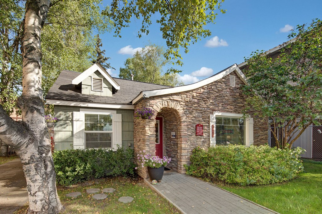 The front office here at Castle Apartments with stone facade and red door, surrounded by lush green trees and bushes. A birch tree and potted flowers add a cozy feel.
