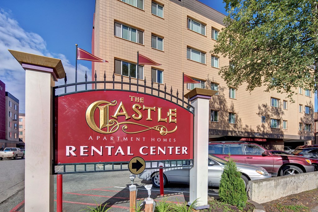 Red sign reading "The Castle Apartment Homes Rental Center" in gold and white stands prominently. Background shows a beige apartment building, parked cars, and a tree, suggesting a residential area.