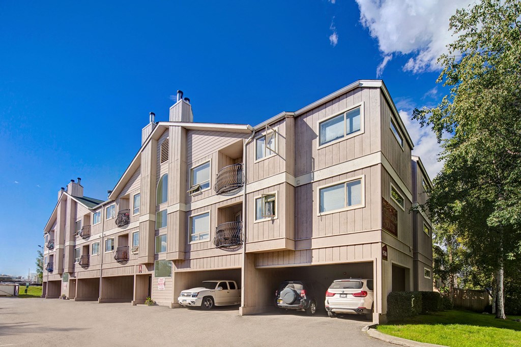 Chugach South Apartments - Exterior Three-story beige Chugach South Apartment building sits under a clear blue sky, with three cars parked in open garages. Trees line the right, creating a serene suburban feel.