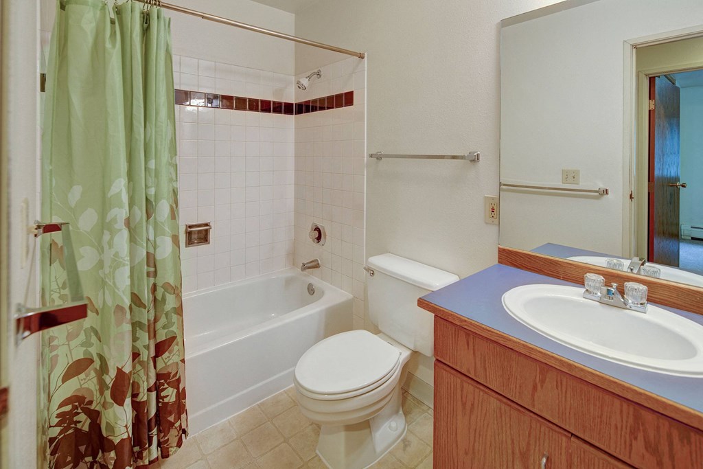 Chugach South Apartments - Bathroom A clean, tidy bathroom here at Chugach South Apartments with a light green shower curtain featuring leaf patterns, a white bathtub, a toilet, and a wooden vanity with a blue countertop.