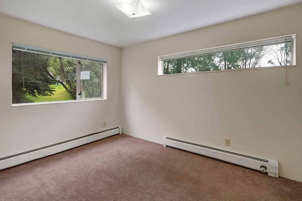 Chugach South Apartments - Bedroom Empty living room here at Chugach South Apartements with beige carpet and cream walls, featuring two large windows showing a green, leafy backyard. The peaceful ambiance is bright and open.