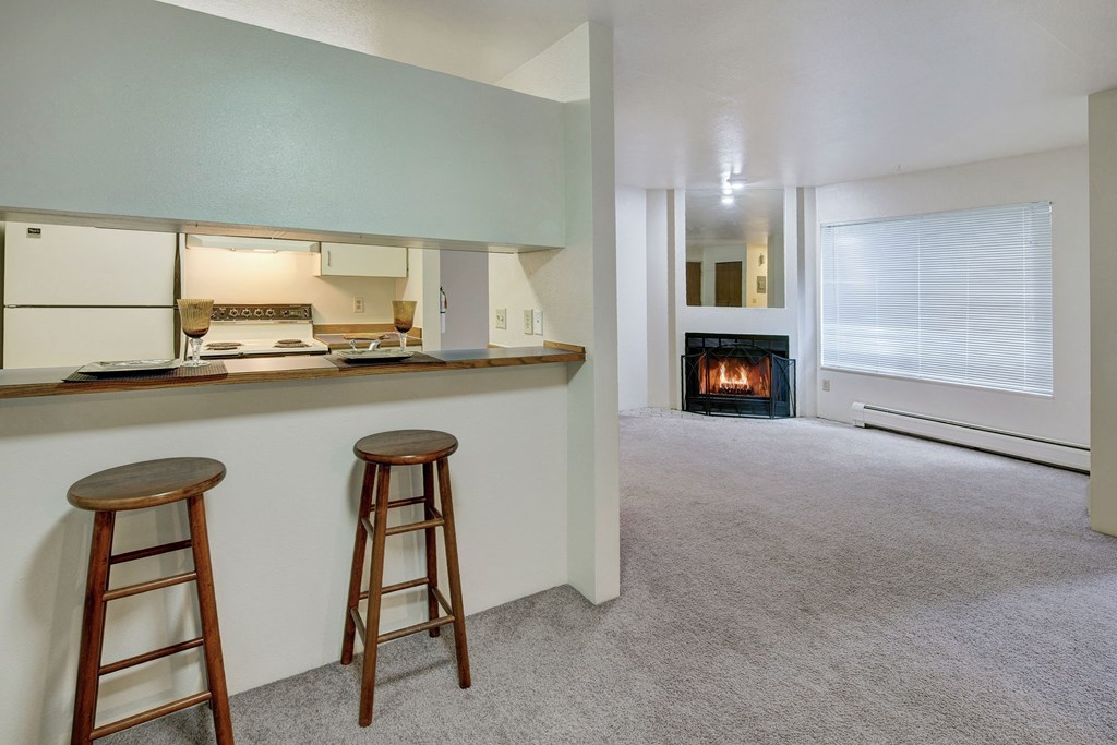 Chugach South Apartments - Dining Area Spacious, cozy living area here at Chugach South Apartments with a grey carpet, featuring a kitchen bar with two wooden stools on the left, and a lit fireplace on the right.