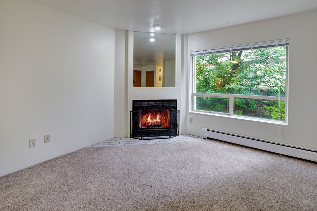Chugach South Apartments - Living Room A cozy room here at Chugach South Apartments with a lit fireplace, beige carpet, and a large window showing green foliage. The atmosphere is warm and inviting, ideal for relaxation.