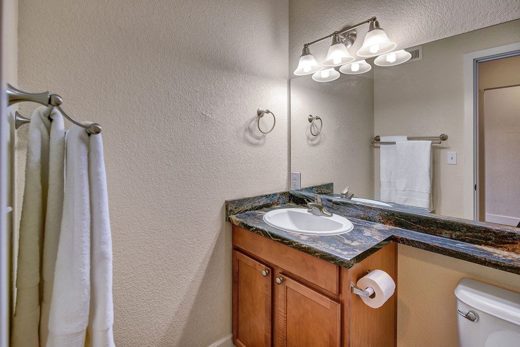 Bathroom here at City View Apartments with wooden vanity, marble countertop, and mirror. Three-light fixture above, white towels on racks, and a roll of toilet paper. Clean and modern.