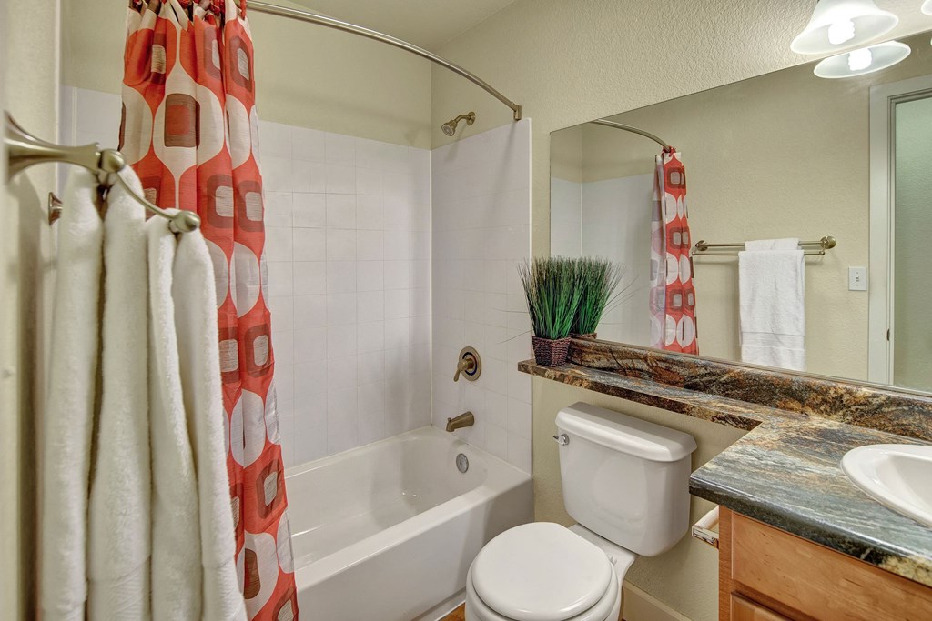 A small bathroom here at City View Apartments with a white tub and colorful red and white shower curtain. A marble countertop with a sink and potted plant adds a touch of elegance.