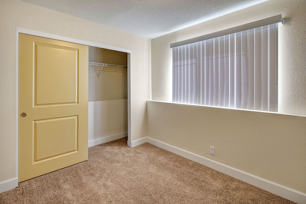 Empty bedroom here at City View Apartments with beige carpet, a partially open sliding yellow closet door on the left, and a bright window with vertical blinds on the right.