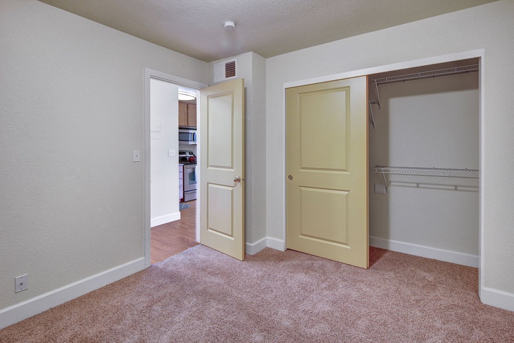Bedroom here at City View Apartments with beige carpet and light walls. A partially open closet with sliding doors is on the right, and an open door leads to a kitchen.