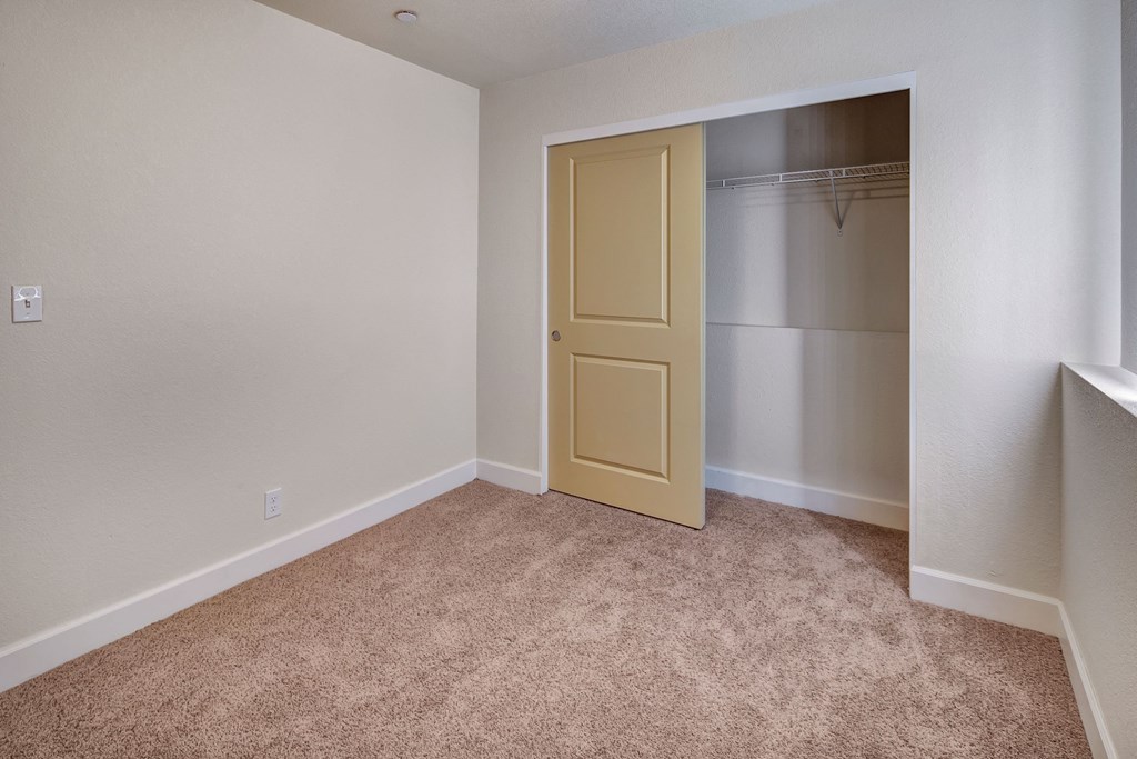 Empty beige bedroom here at City View Apartments with carpet, open closet with a sliding yellow door, white baseboards, and a small window on the right, evoking a clean, neutral feel.