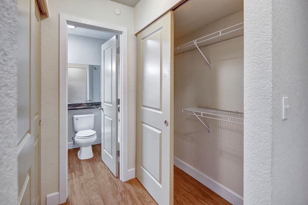 Hallway here at City View Apartments showing an open closet with wire shelves to the right, and a view into a bathroom with a white toilet and mirror on the left. Wood flooring.