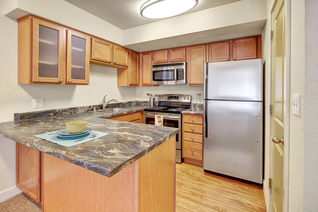 Compact kitchen here at City View Apartments with light wood cabinets and marble countertops. Stainless steel appliances, including a fridge and microwave. Cozy and inviting feel.