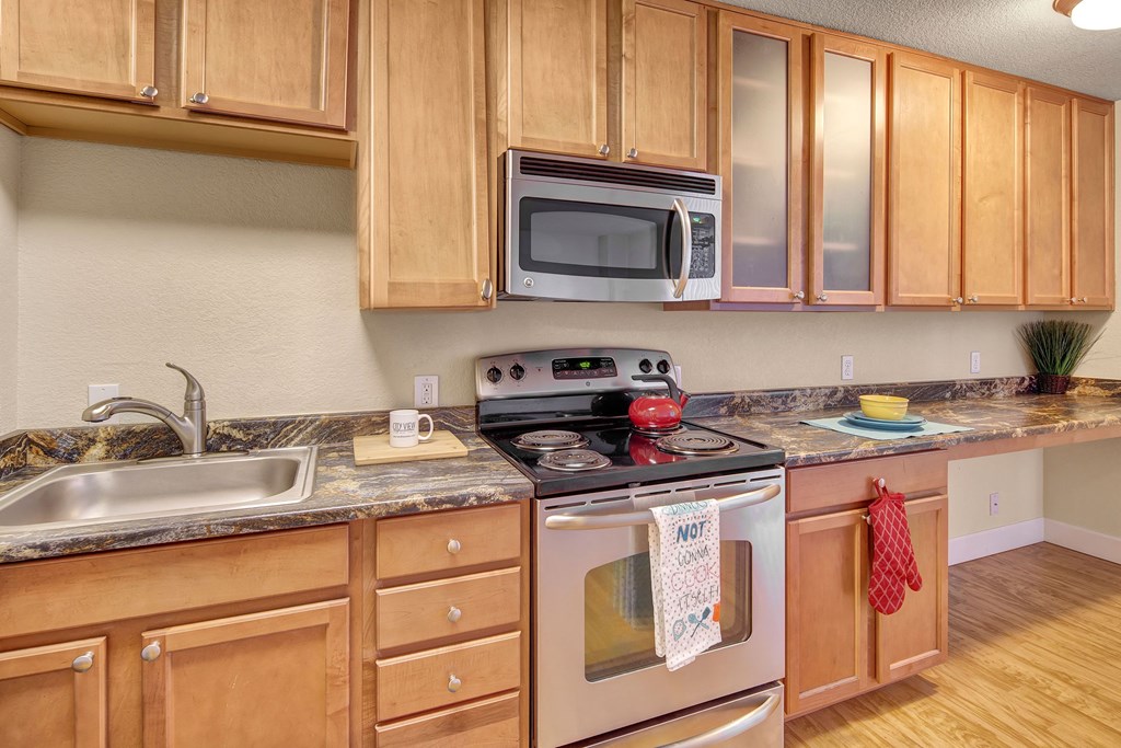 Modern kitchen here at City View Apartments with light wood cabinets, granite countertops, and stainless steel appliances. A red kettle sits on the stove, with oven mitts and towels nearby.