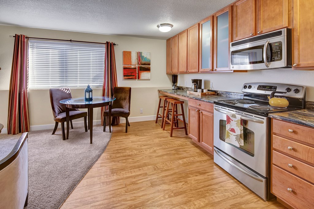 Warm kitchen-dining area here at City View Apartments with wooden cabinets, stainless steel appliances, round glass table, two chairs, large window, and abstract art on the wall.