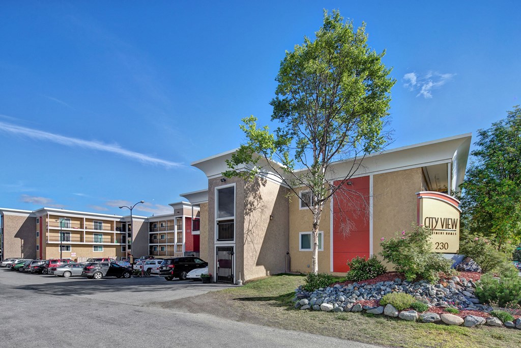 City View Apartment complex under a clear blue sky, featuring modern beige buildings with red accents, a parking lot with cars, and landscaped greenery.