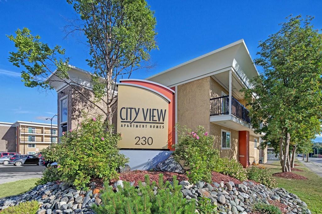 Apartment complex exterior with a sign reading "City View Apartment Homes 230." Surrounded by trees and shrubs, under a clear blue sky. Inviting and serene.