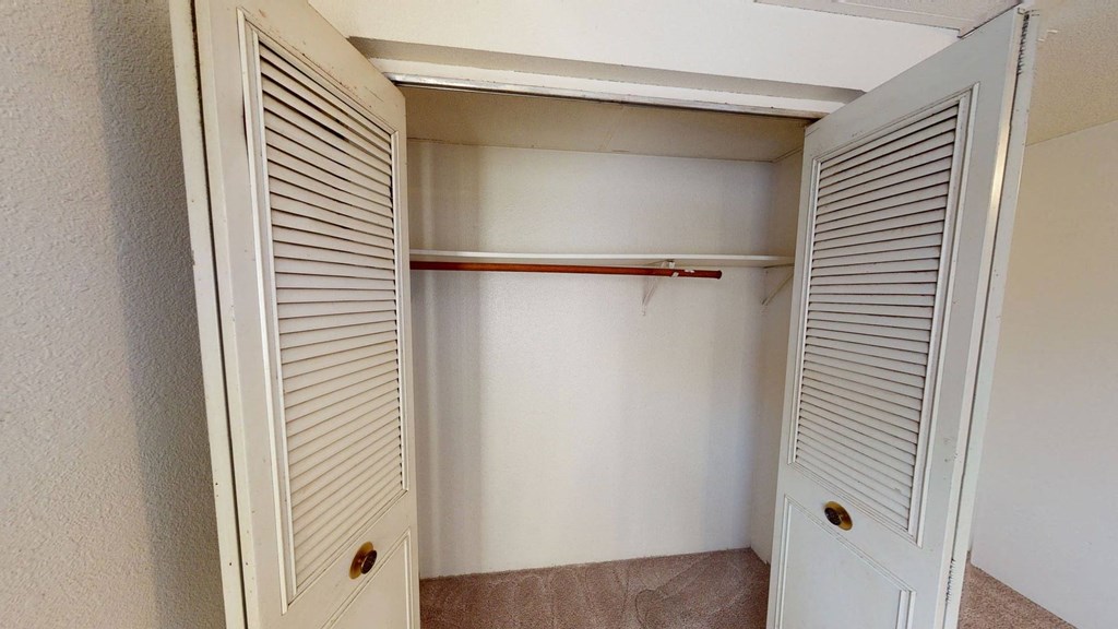 Empty closet here at College View Apartments with open louvered doors, revealing a wooden clothes rod and beige carpet. The ambiance is simple and neutral.