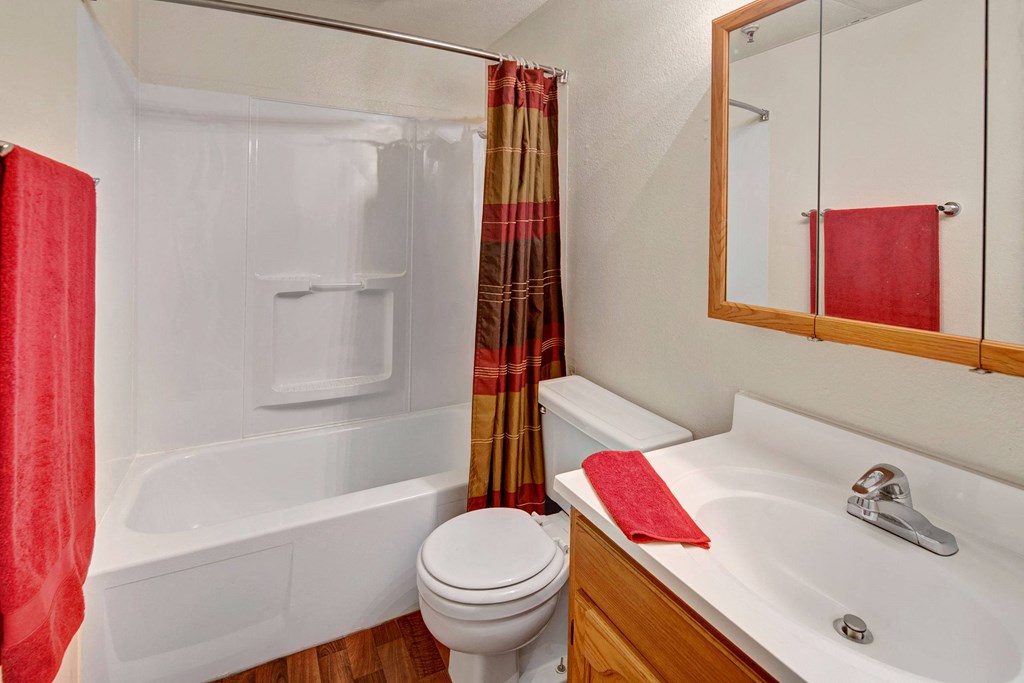 Small bathroom here at College View Apartments with a white bathtub, red and brown shower curtain, red towels, white sink on a wooden vanity, and a mirrored cabinet above.