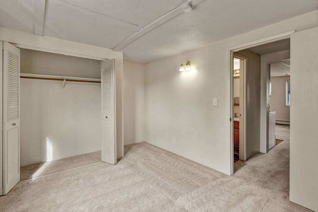 Empty beige carpeted bedroom here at College View Apartments with an open closet on the left, showing hanging rod, and an open door leading to a hallway. Dim wall light creates a cozy feel.
