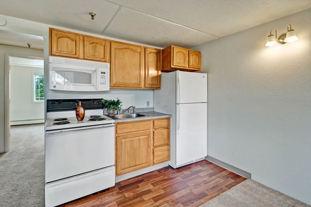 Compact kitchen here at College View Apartments with wooden cabinets, white appliances, and a vase on the stove. Bright room with hardwood floors, adjacent to a carpeted space.