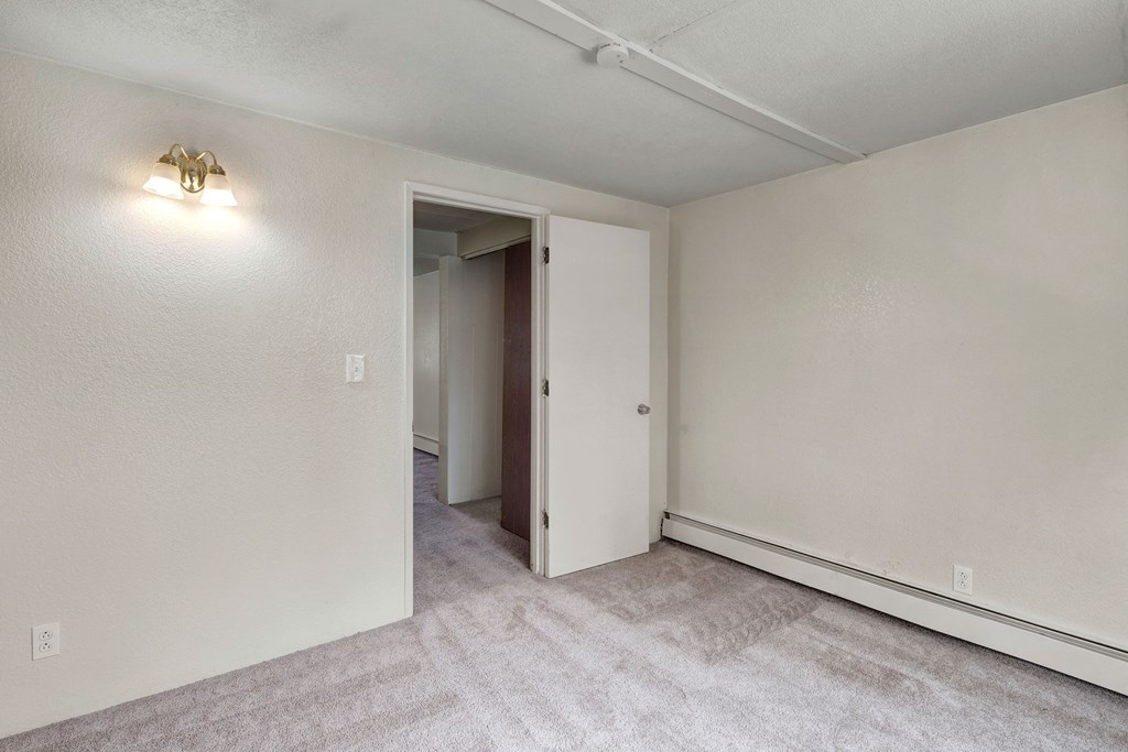 Empty bedroom here at College View Apartments with beige walls, light gray carpet, a closed white door slightly ajar, and a wall sconce casting soft light, creating a neutral tone.