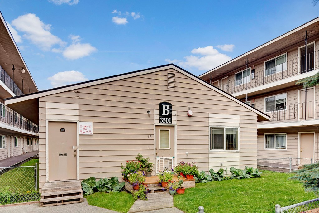 College View Apartments exterior of a beige building labeled "B 3501" with potted plants and flowers in front. Surrounding buildings have balconies. Clear blue sky above.