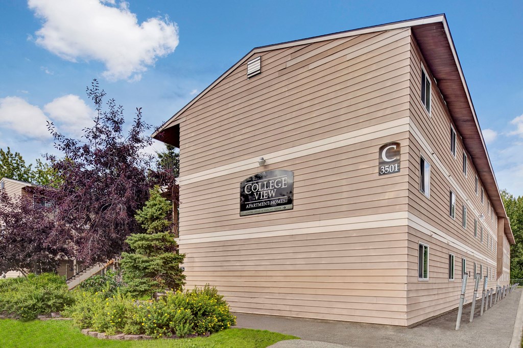 Exterior view of a beige, three-story apartment building with "College View Apartment Homes" sign. Surrounded by green shrubs and a tree under a blue sky.