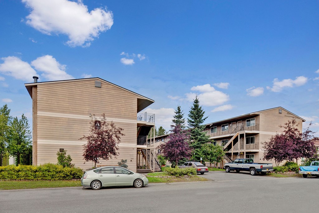 Two beige apartment buildings here at College View Apartments with wooden siding, surrounded by trees and parked cars under a clear blue sky. The scene conveys a peaceful suburban setting.