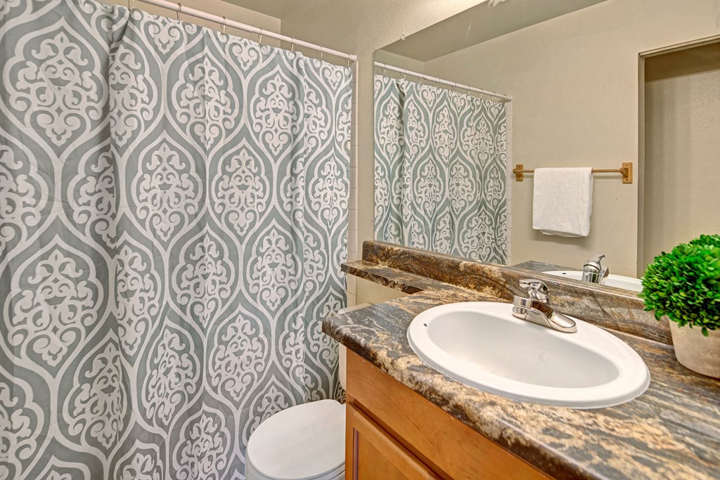 Bathroom here at Conifer Grove Apartments with a decorative green and white patterned shower curtain, a brown marble countertop, green potted plants, and a white towel on a rack.