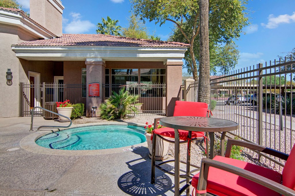 Small hot tub in a sunlit courtyard here at Dobson Towne Centre Apartments, surrounded by a brown fence and a building. Red chairs and table add a vibrant touch. Relaxing atmosphere.