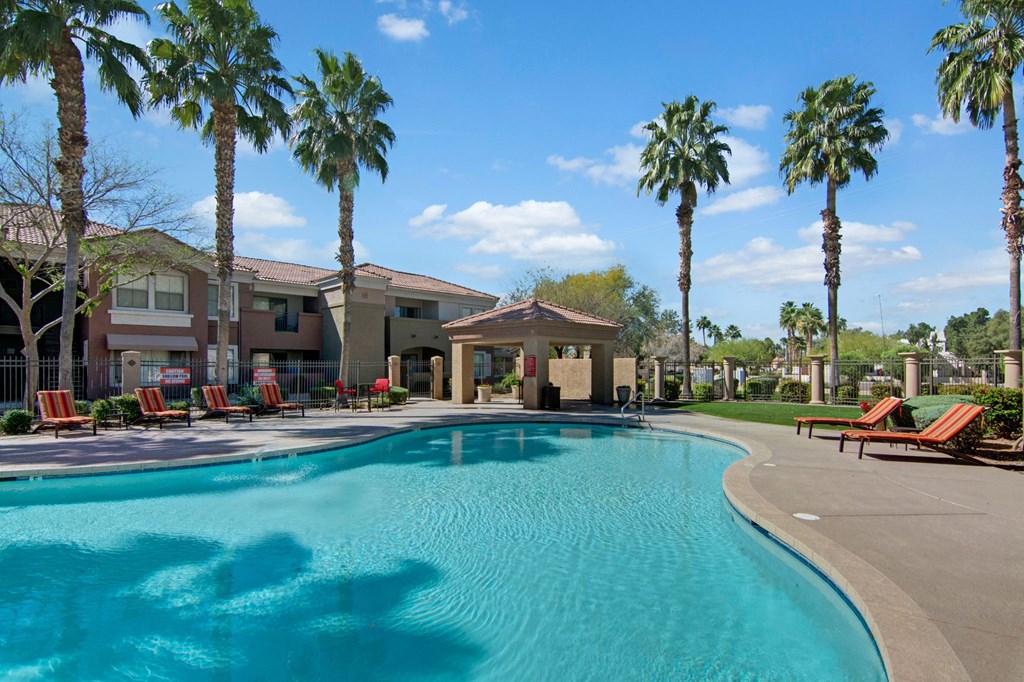 Outdoor pool area here at Dobson Towne Centre Apartments with clear blue water and red-striped lounge chairs lined up on the deck. Tall palm trees and a building are in the background under a clear blue sky.