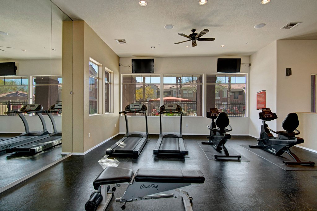 A bright gym here at Dobson Towne Centre Apartments with two treadmills and two recumbent bikes facing large windows. A weight bench is in the foreground; the atmosphere is clean and inviting.