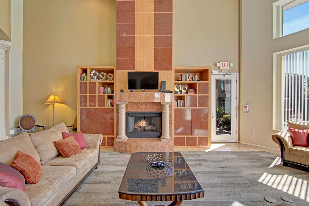 Spacious resident lounge here at Dobson Towne Centre Apartments with beige walls, tall fireplace, and wooden shelves. Cozy beige sofa with red cushions. Sunlight streams through large windows. Relaxed, inviting ambiance.