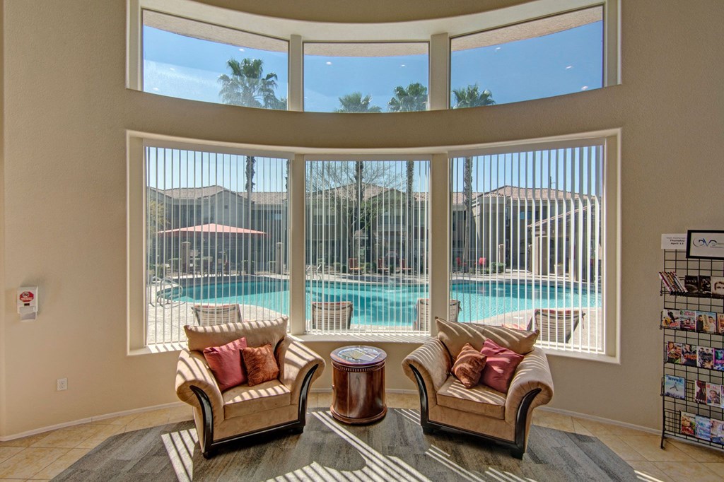 Cozy seating area here at Dobson Towne Centre Apartments with two beige armchairs and a round table, facing large windows overlooking a sunny pool, creating a relaxed and inviting atmosphere.