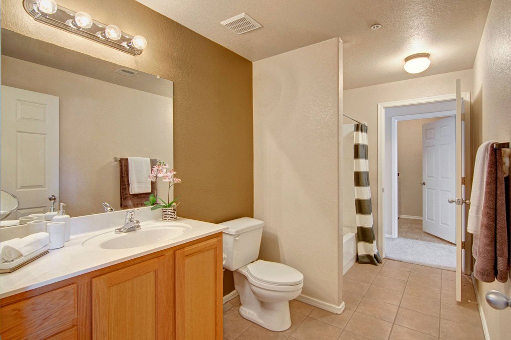 Bright bathroom here at Dobson Towne Centre Apartments with a white sink on a wooden cabinet, large mirror, and overhead lights. A toilet and shower with a striped curtain are visible.